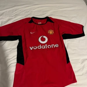 Röd Manchester United fotbollströja - Säljer en klassisk röd Manchester United fotbollströja från Nike med korta ärmar. Tröjan har svart och vit detaljer samt sponsorlogga på framsidan. På baksidan finns spelarnamnet och nummer 7. Perfekt för fans av laget!