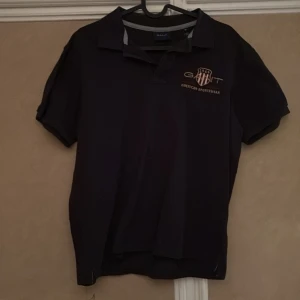 Mörkblå pikétröja från GANT - Säljer en stilren mörkblå pikétröja från GANT med korta ärmar och klassisk krage. Tröjan har en broderad logotyp i guld på bröstet. Perfekt för en avslappnad men ändå snygg look.