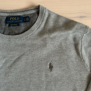 Grå tröja från Ralph Lauren - Säljer en stilren grå tröja från Ralph Lauren i mjuk pima bomull. Tröjan har en broderad logotyp på bröstet och ribbade muddar vid ärmslut och nederkant. Perfekt för en avslappnad och snygg look.