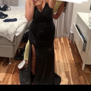 Svart glittrig långklänning - Elegant svart långklänning med glittrande detaljer och en djup v-ringning. Klänningen har en slits framtill och snörning i ryggen för en perfekt passform. Perfekt för en glamorös kväll. Använd bara en gång 