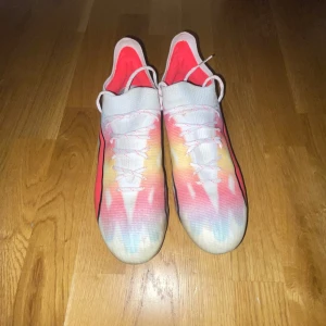 Färgglada fotbollsskor från Puma - Säljer ett par färgglada fotbollsskor från Puma med en gradient av rosa, gul och blå. Skorna har snörning och en strömlinjeformad design med en svart Puma-logga på sidan. Perfekta för att sticka ut på planen!