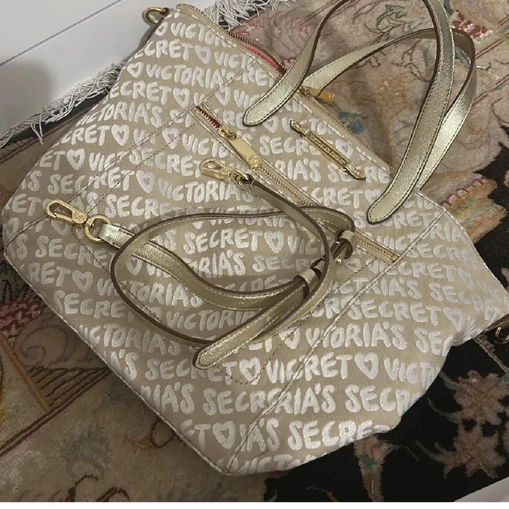 Snygg beige axelväska från Victoria's Secret med ett mönster av vita hjärtan och text. Väskan har guldfärgade detaljer och dragkedja. Perfekt för att bära med sig det viktigaste med stil.. Laukut & Käsilaukut.