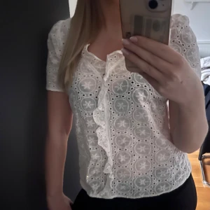 Vit blus med spetsdetaljer - Säljer en söt vit blus med spetsdetaljer och korta ärmar. Blusen har en vacker volang framtill och ett fint broderat mönster. Perfekt för en somrig look!