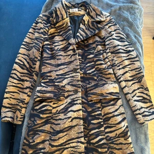 Tigermönstrad kappa från Pikol - Säljer en unik tigermönstrad kappa från Pikol. Perfekt vår kappa! Den har en klassisk krage och knappar framtill. Perfekt för att sticka ut i mängden med sitt djärva mönster. One of a kind! 