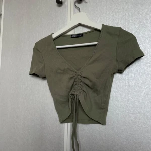 Olivgrön ribbad topp från Zara - Säljer en olivgrön ribbad topp från Zara med korta ärmar och v-ringning. Toppen har en snygg knytdetalj framtill som ger en trendig look. Perfekt för en stilren outfit!