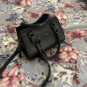 Svart axelväska med dragkedja - Snygg svart axelväska i syntetmaterial med stilrena detaljer. Väskan har en praktisk dragkedja och dubbla handtag för enkel bärning. Perfekt för att hålla dina saker organiserade och säkra. En tidlös accessoar som passar till många tillfällen.