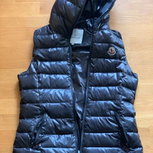 Svart dunväst från Moncler - Snygg svart dunväst från Moncler med dragkedja och huva. Perfekt för kyligare dagar när du vill hålla stilen. Västen har en quiltad design och två praktiska fickor med dragkedja.