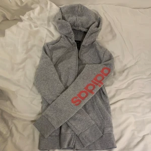Grå hoodie / kofta från Adidas - Säljer en grå kofta från Adidas med rosa logga på ärmen. Tröjan har en dragkedja framtill och en bekväm huva. 