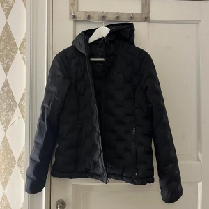 Svart quiltad jacka med huva - Snygg svart quiltad jacka med huva och dragkedja framtill. Jackan har långa ärmar och två praktiska fickor med dragkedja. Perfekt för kyligare dagar.