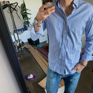 Blå och vit randig skjorta från Ralph Lauren - Snygg blå och vit randig skjorta från Ralph Lauren med en gul broderad logga på bröstet. Skjortan har långa ärmar och klassisk krage. Perfekt för en stilren look. Passar mer som en M
