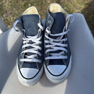 Blå sneakers från Converse - Säljer ett par klassiska blå Converse All Star sneakers med vita snören och tåhätta. Skorna har den ikoniska loggan på sidan och en bekväm passform. Perfekta för en avslappnad stil.