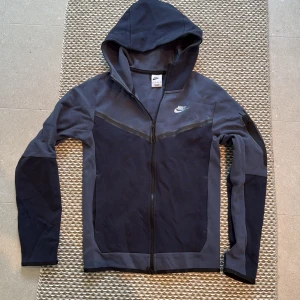 Grå och svart hoodie från Nike - Säljer en snygg grå och svart hoodie från Nike med dragkedja och huva. Jackan har långa ärmar och en stilren design med Nike-logga på bröstet. Perfekt för en sportig look!