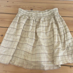 Beige rutig kjol - Charmig beige kjol med rutigt mönster. Den har en bekväm elastisk midja och en luftig passform som ger en avslappnad stil. Perfekt för varma dagar.