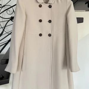 Beige kappa med knappar - Elegant beige kappa med dubbelknäppning och långa ärmar. Perfekt för höst och vår. Klassisk design med stilrena detaljer. Toppen kvalitet! 