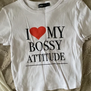 Vit t-shirt med tryck från FB Sister - Vit t-shirt från FB Sister med svart text och ett rött hjärta på bröstet där det står 'I ❤️ MY BOSSY ATTITUDE'. Klassisk rund hals och korta ärmar. Perfekt statement-plagg för dig som gillar att sticka ut.