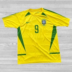 Retro Brasilien fotbollströja (Ronaldo 9) - Säljer en gul Brasilien fotbollströja från Nike med gröna detaljer och nummer 9 på framsidan. Tröjan har korta ärmar och är tillverkad i Dri-FIT material för optimal komfort. Perfekt för fotbollsfans som vill visa sitt stöd för laget!