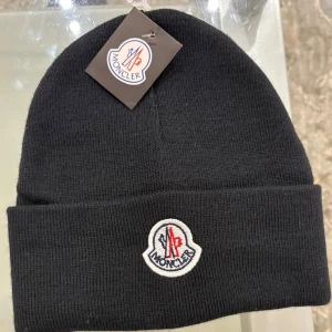 Moncler mössa - Snygg svart mössa från Moncler med broderad logotyp framtill. Perfekt för att hålla värmen under kyliga dagar. Stilren design som passar till det mesta.