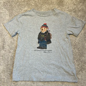Grå Ralph Lauren t-shirt med Polo Bear på framsidan - Säljer en grå t-shirt från Ralph Lauren med en cool Polo Bear-tryck på framsidan. Björnen har på sig en vinteroutfit med mössa och jacka. Perfekt för en avslappnad stil!