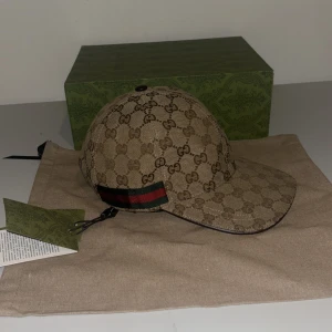 Beige keps från Gucci - Snygg beige keps från Gucci med det ikoniska GG-mönstret. Kepsen har en justerbar rem med röd och grön rand på baksidan. Perfekt för att ge en lyxig touch till din outfit.