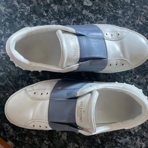 Valentino Garavani sneakers i vitt och blått - Snygga Valentino Garavani sneakers i vitt läder med en bred blå/grå rem över ovansidan. Skorna har en rund tå och en unik design med pyramidformade detaljer på hälen. Perfekta för att ge din outfit en lyxig touch.