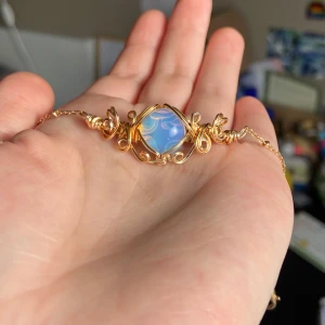 Guldarmband med blå sten ”opalit” - Elegant armband i guld med en vacker blå sten i mitten. Armbandet har detaljerade guldfärgade snirklar som ramar in stenen och ger en unik look. Perfekt för att ge en touch av stil till din outfit.