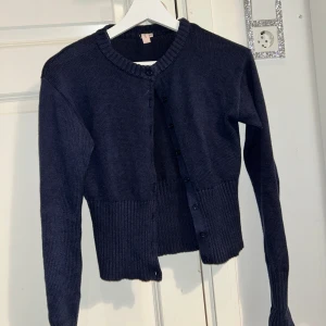 Mörkblå kofta med knappar från Nelly - Säljer en stilren mörkblå kofta med knappar framtill. Den har en ribbad kant vid ärmslut och nederkant, vilket ger en snygg passform. Perfekt för kyligare dagar! Använd några gånger men annars i super skick!