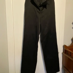 Svarta satin kostymbyxor från H&M - Snygga svarta kostymbyxor från H&M med hög midja och raka ben. Byxorna har en stilren design med diskreta fickor baktill och stängs med dragkedja och knapp framtill. Perfekta för en elegant look. Ena fållen släppt på ena benet, men enkelt att åtgärda. 