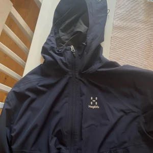 Marin blå vindjacka från Haglöfs - Säljer en marin blå vindjacka från Haglöfs med huva och dragkedja. Perfekt för utomhusaktiviteter med sin funktionella design och stilrena look. Jackan har en diskret logga på bröstet och är tillverkad i ett lätt och smidigt material. Köpt för 1300kr men säljer den nu för bara 500kr
