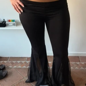 Svarta byxor med transparenta detaljer - Snygga svarta byxor med låg midja och transparenta detaljer vid benen. Byxorna har en tight passform upptill och vida ben nedtill, vilket ger en trendig look. Perfekta för en kväll ute eller en speciell tillställning.