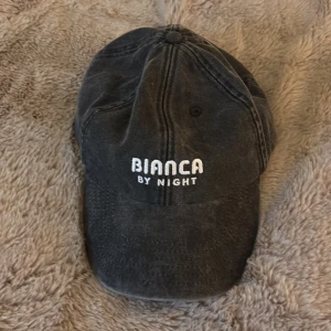 Svart keps från Bianca By Night - Snygg svart keps med broderad text 'BIANCA BY NIGHT' framtill och 'Coca-Cola' baktill. Justerbar rem med metallspänne för perfekt passform.