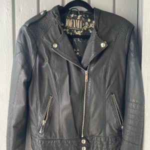 Svart skinnjacka från Jofama - Snygg svart skinnjacka från Jofama med coola detaljer som dragkedjor och knappar. Jackan har en klassisk bikerstil med quiltade axlar och en blommig insida. Ny! Aldrig använd. Stl 40