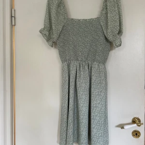 Grön blommig klänning från NA-KD - Säljer en söt grön klänning från NA-KD med små vita blommor. Klänningen har puffiga kortärmar och en smockad överdel som ger en fin passform. Perfekt för vår och sommar!