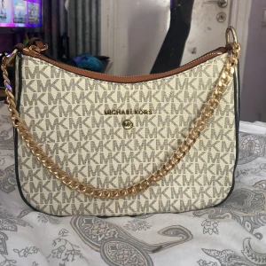 Beige axelväska från Michael Kors - Snygg beige axelväska från Michael Kors med det klassiska MK-mönstret. Väskan har en guldkedja och en justerbar axelrem i brunt läder. Den stängs med dragkedja och har en praktisk innerficka. Perfekt för att ge din outfit en lyxig touch.