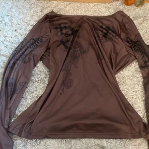 Brun långärmad topp med tryck - Säljer en brun långärmad topp med unikt tryck av fåglar och mönster. Perfekt för dig som vill ha något annorlunda i garderoben. Toppen har en lös passform och är tillverkad i ett mjukt material.