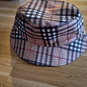 Rutig bucket hat - Snygg bucket hat med klassiskt rutigt mönster i beige, svart och rött. Perfekt för en stilren look oavsett väder. Hatten har en bred kant och är tillverkad i ett lätt material.