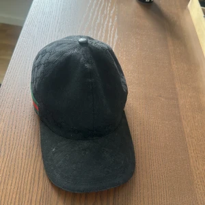 Svart keps från Gucci - Snygg svart keps från Gucci med diskret mönster och klassiska gröna och röda ränder på sidan. Kepsen har en justerbar rem baktill för perfekt passform. Perfekt för att ge din outfit en lyxig touch.