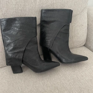 Svarta stövlar från Tiger of Sweden - Snygga svarta stövlar i skinn från Tiger of Sweden med krokodilpräglat mönster. De har en spetsig tå och en stabil klack som ger en elegant look. Perfekta för att lyfta vilken outfit som helst. De går att använda både upp-och nedvikta. OBS. Uppklippta i ett par sömmar då de var svinhårda, det är dock ingenting som märks när man använder de nedvikta. Därav priset. Välanvända och supersköna!