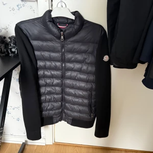 Svart quiltad jacka från Moncler - Säljer en stilren svart quiltad jacka från Moncler. Jackan har långa ärmar och dragkedja framtill. Den är perfekt för kyligare dagar och har en klassisk design med Moncler-logga på ärmen.