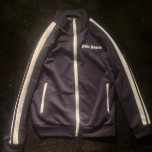 Mörkblå track jacket från Palm Angels - Säljer en snygg mörkblå tröja  från Palm Angels med vita ränder längs ärmarna och dragkedja framtill. Tröjan har en hög krage och två fickor med dragkedjor. Perfekt!