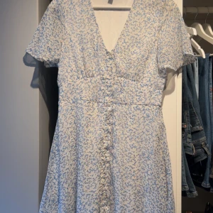 Blommig klänning med knappar - Söt blommig klänning i ljusblått och vitt med v-ringning och korta ärmar. Klänningen har knappar framtill och en tajtare passform i midjan.