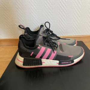 Adidas NMD sneakers i grått och rosa - Snygga Adidas NMD sneakers i grått med rosa detaljer och svarta snören. Skorna har en modern design med den ikoniska vita sulan och rosa ränder på sidan. Perfekta för en sportig look.