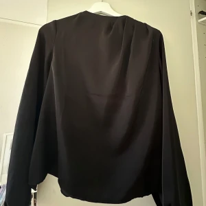 Svart satinblus från Kappahl - Elegant svart blus med långärmad design. Perfekt för en stilren look. Passar bra till både formella och avslappnade tillfällen. 