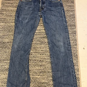 Levis 501 jeans - Klassiska Levis 501 jeans i blå denim med knappgylf. Dessa jeans har en straight passform och är tillverkade i slitstarkt jeanstyg. Perfekta för en tidlös stil.