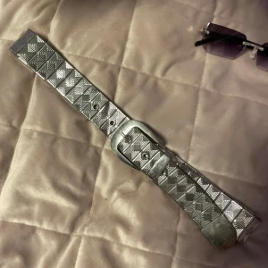 Silvrigt metalliskt bälte med rutigt mönster - Snyggt silvrigt metalliskt bälte med ett unikt rutigt mönster. Bältet har en klassisk spänne och flera hål för justering. Perfekt för att ge en glittrig touch till din outfit. Omkretsen med längsta hålet är ungefär 80 cm!  