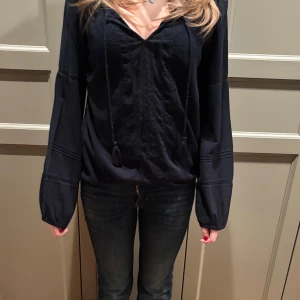 Svart blus med v-ringning från Abercrombie and Fitch  - Säljer en elegant svart blus med v-ringning och långa ärmar. Blusen har fina detaljer med knytband och broderier framtill. Perfekt för en stilren look.