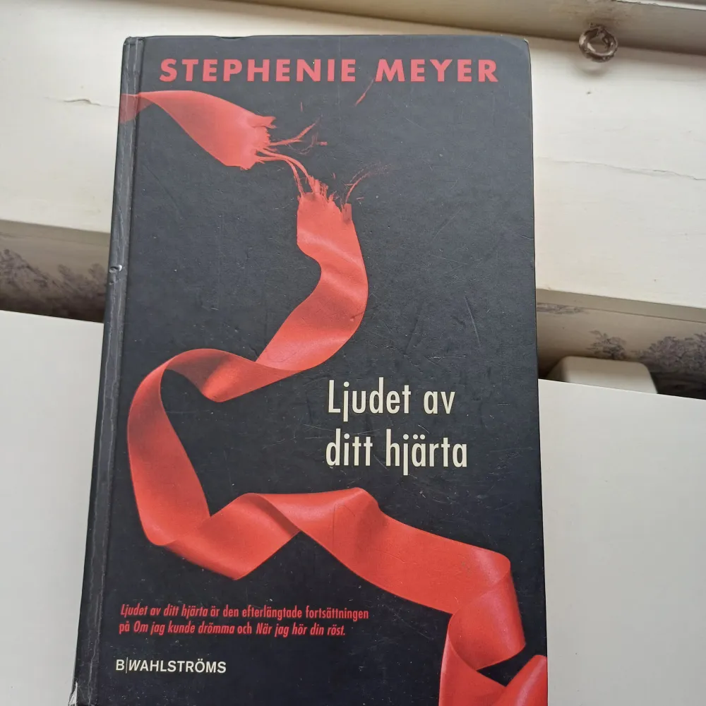 Boken 'Ljudet av ditt hjärta' av Stephenie Meyer är en spännande fortsättning i en populär serie. Den har ett dramatiskt omslag med en röd banderoll mot en svart bakgrund. Perfekt för dig som älskar romantik och övernaturliga berättelser.. Böcker.