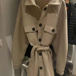Beige kappa med bälte - Snygg beige kappa med knappar och bälte i midjan. Den har långa ärmar och två stora fickor framtill. Perfekt för att hålla stilen i kyligare väder. Storlek S men oversized 