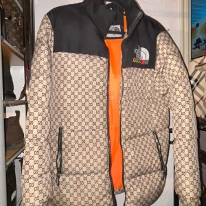 Beige och svart pufferjacka från The North Face x Gucci - Säljer en snygg beige och svart pufferjacka från The North Face x Gucci med ett ikoniskt mönster. Jackan har en dragkedja och är långärmad med en cool orange insida. Perfekt för att hålla stilen i kyligare väder. Priset går att diskutera då jag vill få bort de snabbt då jag håller på flytta 