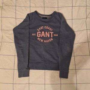 Blå sweatshirt från GANT💙 - Säljer en snygg blå sweatshirt från GANT med tryck 'East Coast New Haven' i rosa på framsidan. Tröjan har långa ärmar och ribbade muddar vid ärmslut och nederkant. Perfekt för en avslappnad stil.