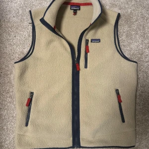 Beige fleeceväst från Patagonia - Säljer en beige fleeceväst från Patagonia med dragkedja och kontrasterande mörkblå detaljer. Västen har två fickor med dragkedjor och en hög krage. Perfekt för lager-på-lager under kyliga dagar.